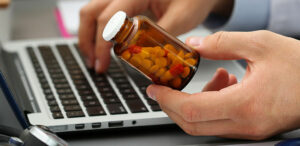 Man holding a glass pill jar near a laptop.