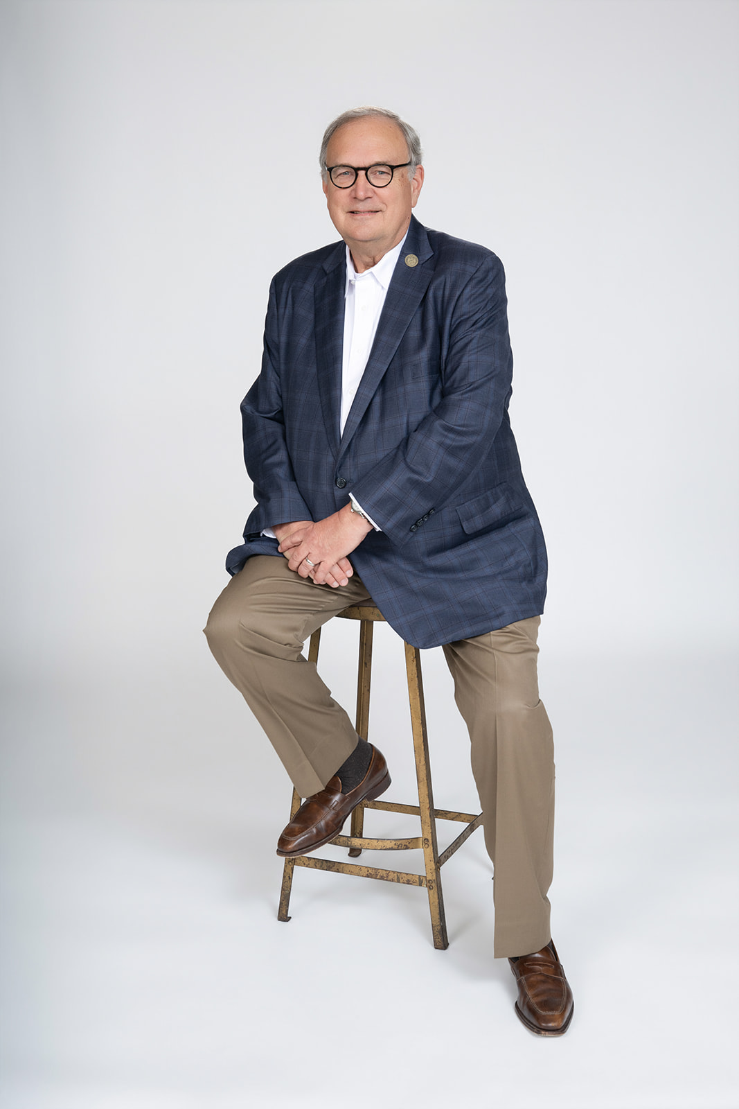 Photo of Warren Erdman wearing a blue plaid sport coat, sitting on a stool in a photography studio with white background