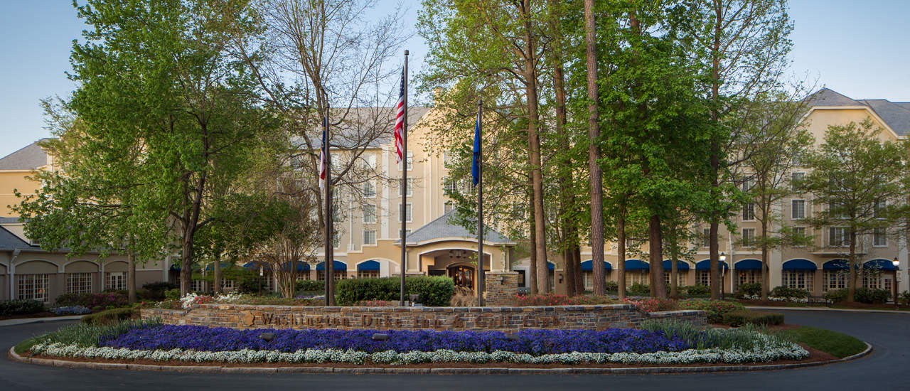 Exterior image of the Washington Duke Inn main entrance and circle drive with mature trees and blue flowers in landscape.