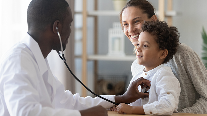 A young boy sits on his mother's lap while a doctor uses a stethoscope to listen to the boy's heart.